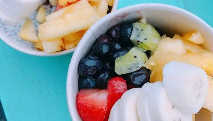 Two bowls of mixed fruit are placed on a teal surface, reminiscent of the vibrant smoothies and acai bowls in Kauai. The front bowl contains sliced bananas, strawberries, blueberries, kiwi, and pineapple chunks. The back bowl has a blend of pineapple pieces and what appears to be a chia seed pudding.