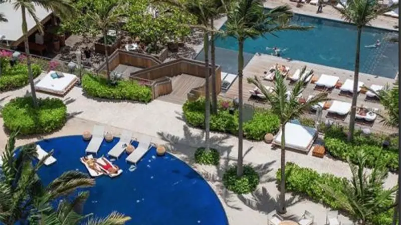 A tropical resort features two swimming pools surrounded by lush greenery and palm trees. One pool is circular with several lounge chairs and umbrellas, while the other is rectangular with additional sunbeds and a wooden deck area. Guests are relaxing and swimming.