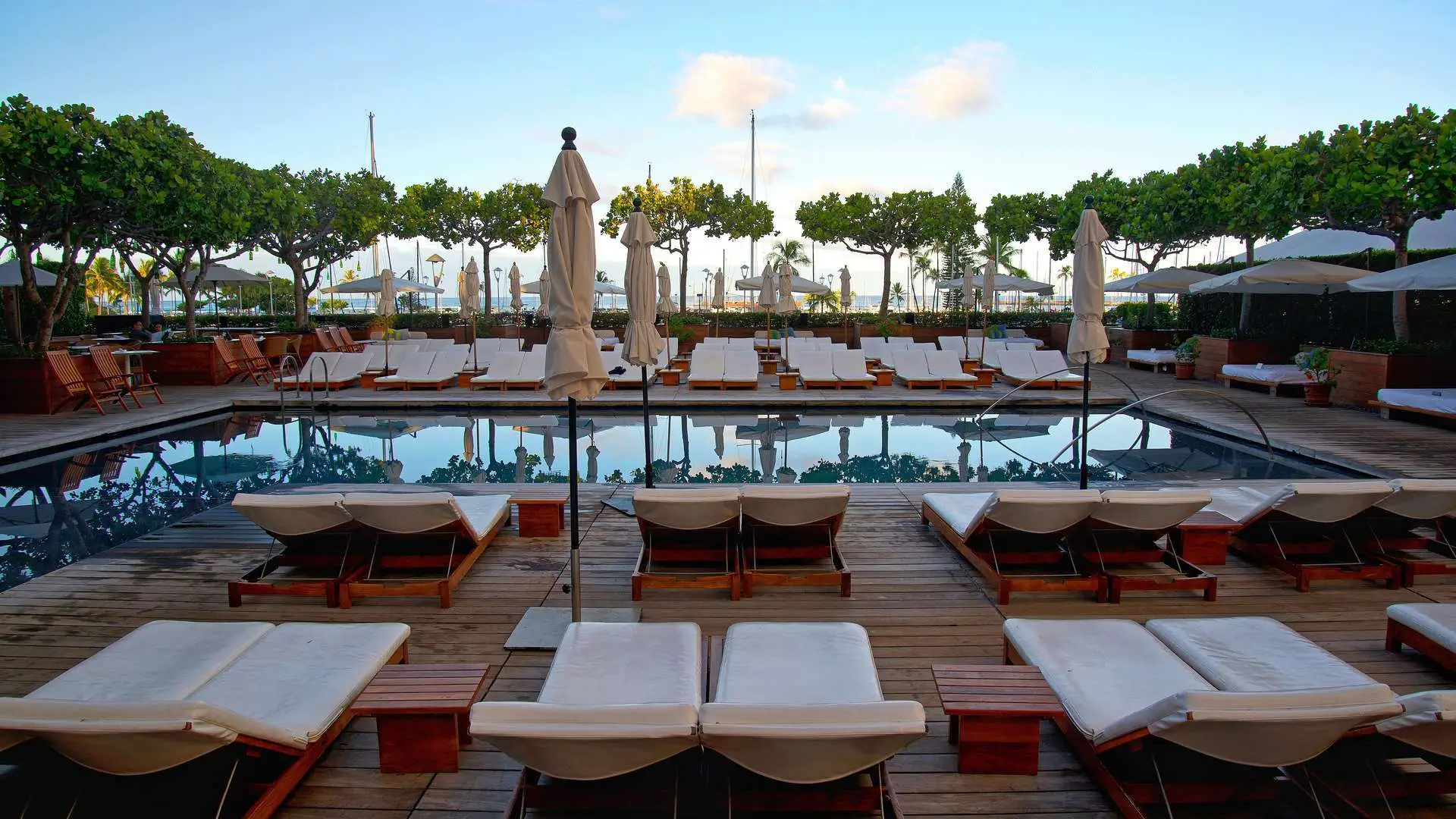 A serene pool area with numerous white cushioned lounge chairs and umbrellas arranged around a rectangular pool. Trees and greenery surround the area, with boats and masts visible in the background under a clear sky—perfect for idyllic group getaways in Oahu.