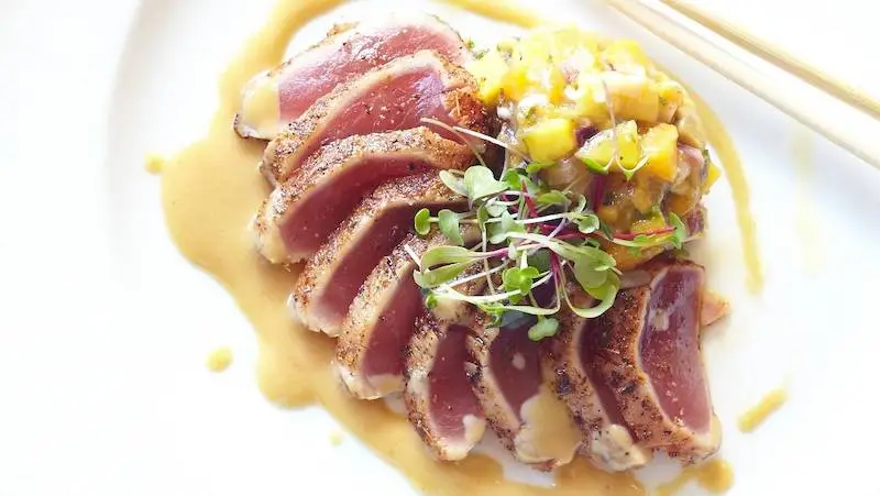 A gourmet dish featuring several slices of seared tuna arranged in a fan shape on a white plate. The tuna, lightly seared with a pepper crust, is drizzled with creamy sauce and garnished with microgreens. Served alongside mango salsa, it's the best gluten-free delicacy in Wailea. Chopsticks are visible.