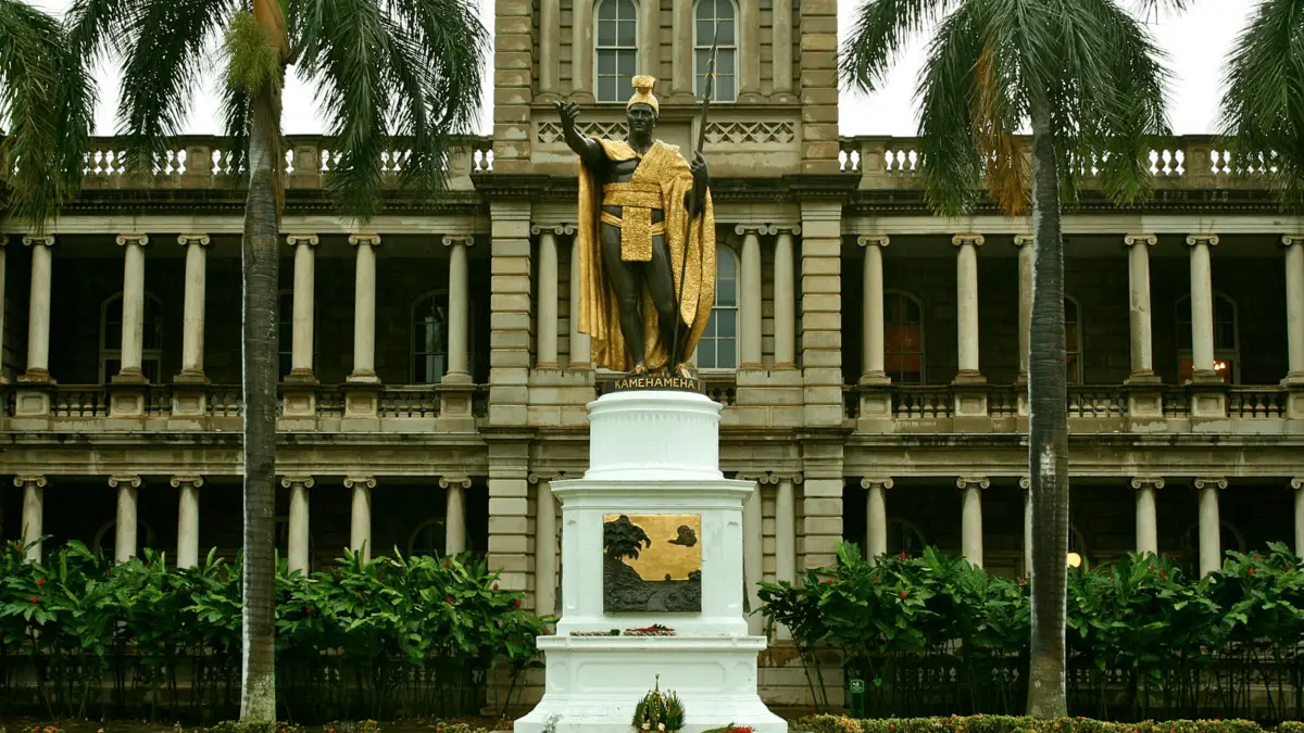 A bronze statue of King Kamehameha I adorned with a golden robe and headdress stands prominently on a white pedestal. This iconic monument, rich in Hawaiian history, is situated in front of a historic building, with tall palm trees and lush greenery reminiscent of a traditional Hawaiian luau.