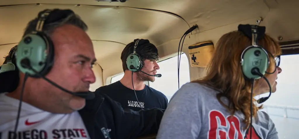 Three people wearing headsets sit inside a small aircraft, looking out the windows. The person on the left wears a San Diego State shirt, the person in the middle gazes straight ahead, and the person on the right has long hair and looks out at their Seaplane Adventures.