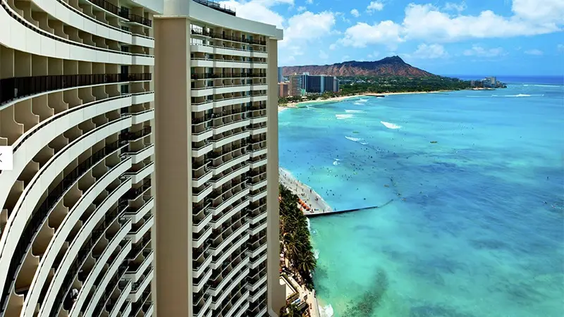 A high-rise building with curved balconies overlooks a turquoise ocean. The shoreline is dotted with people, and the background features a distant mountain range under a partly cloudy blue sky.