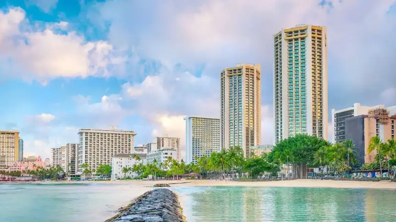 A scenic view of a beach in Oahu with a calm shoreline and a rock groyne extending into the water. Several high-rise buildings and palm trees line the background against a blue sky with scattered clouds. Ideal for group getaways, the city's skyline enhances this tropical, urban beach atmosphere.