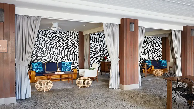 An outdoor seating area featuring wooden furniture with blue and white cushions, surrounded by modern decor. Curtains are drawn to create semi-private spaces. The floor is concrete, and the walls have a bold black and white abstract pattern. Two wicker stools are visible.
