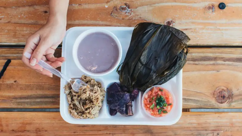 A person is holding a spoon over a tray of food. The tray includes a wrapped tamale, a bowl of purple atole, shredded meat, a portion of purple potatoes, and a small bowl of pico de gallo. The vibrant spread on the wooden table evokes the rich tapestry of Hawaiian luau history.