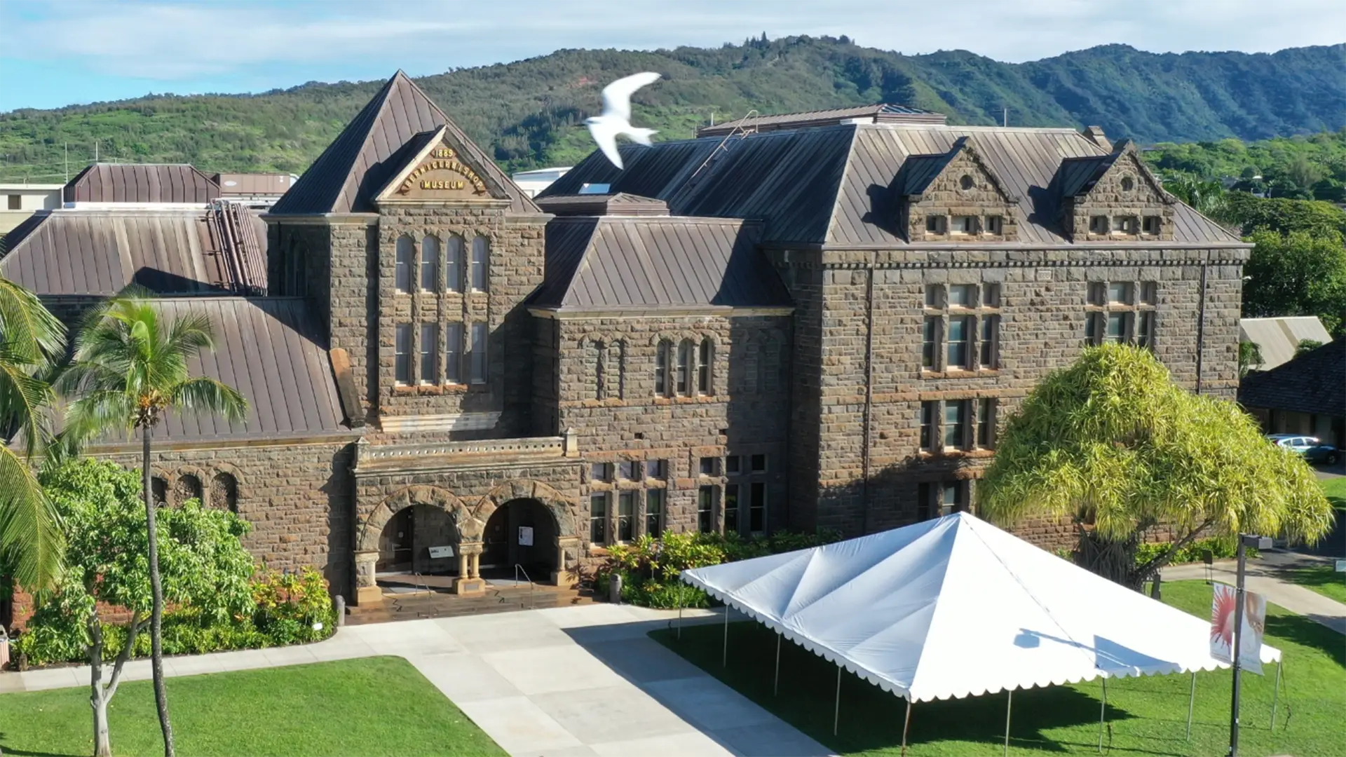 A historic stone building with a steeply pitched roof and arched entrances stands surrounded by grass, palm trees, and a large white tent. A seagull flies above the building, and green hills rise in the background under a clear blue sky.