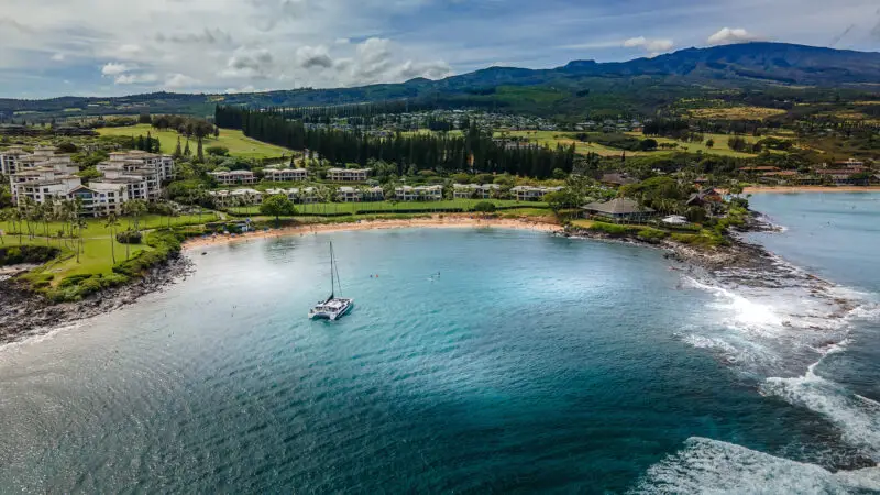 Aerial view of a serene bay on Maui with a sailboat anchored near the shore, surrounded by lush greenery and buildings nestled along the coastline. Rolling hills and mountains form the backdrop under a partially cloudy sky. The turquoise water of the bay is calm and inviting, perfect for family beaches.