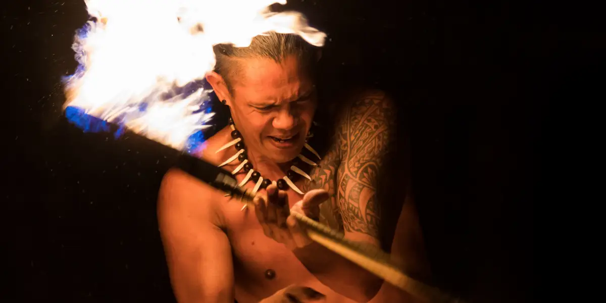 A person with a tribal tattoo on their arm and a necklace made of teeth or claws is performing a fire dance, reminiscent of Hawaiian luau history. They hold a lit torch with intense concentration, the dark background emphasizing the flame above their head and their focused expression.