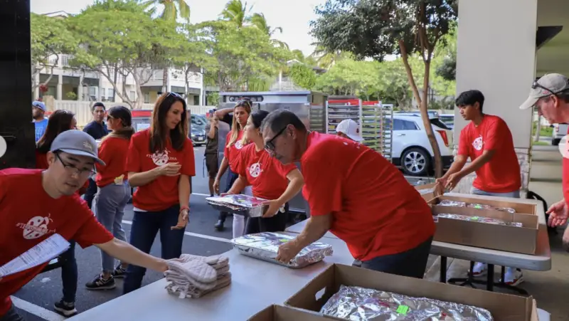 A group of volunteers wearing red shirts work together under a clear sky at one of the Ko Olina Annual Events. They are organizing and packing trays of food. Some handle paperwork while others arrange the trays on tables. Trees and buildings can be seen in the background.