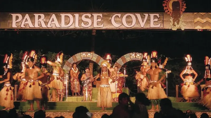 A group of performers in traditional Polynesian attire, including grass skirts and headdresses, are dancing on a stage under a sign that reads "Paradise Cove." The background is dark, highlighting the well-lit performers. This vibrant display is one of the cherished highlights at Ko Olina Annual Events.