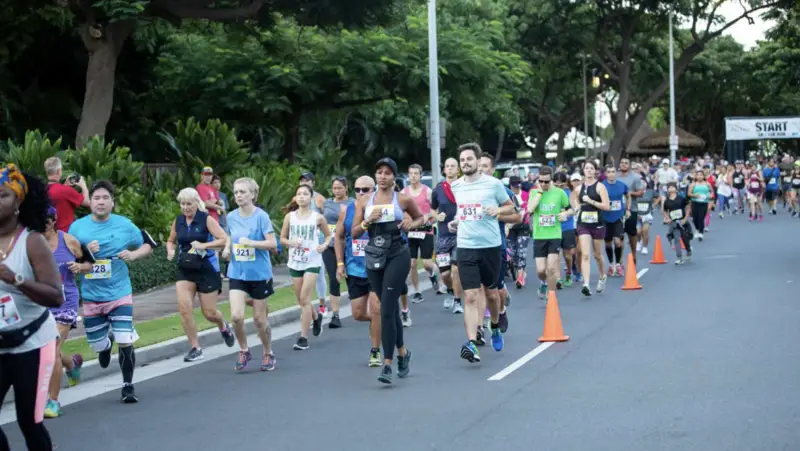 A large group of runners, including people of different ages and genders, participates in the Ko Olina Annual Events race on a paved road lined with orange cones. Trees and greenery surround the race area, and a "START" banner is visible in the background.