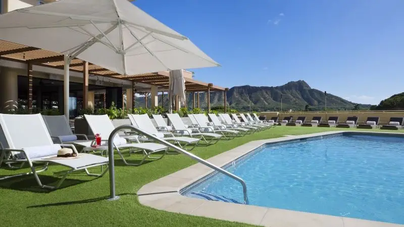 A pristine outdoor pool surrounded by white lounge chairs, some with personal items like hats and drinks. A large white sunshade umbrella stands nearby. In the background, a patio with wooden pergolas and scenic mountains under a clear blue sky are visible.
