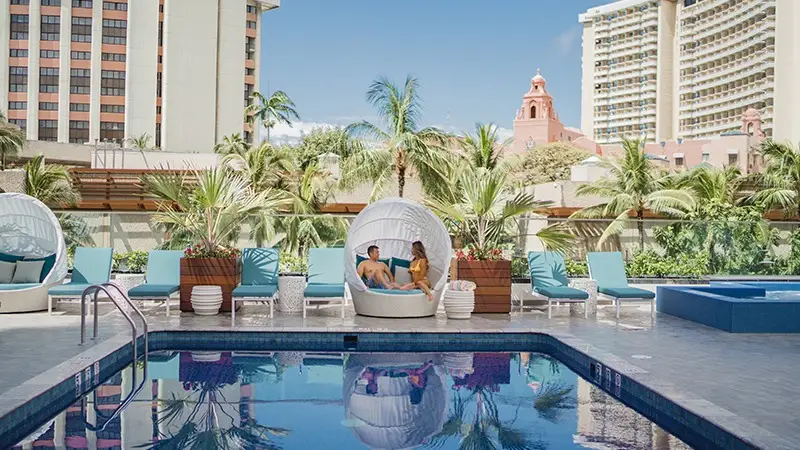 A serene outdoor pool area at a hotel featuring lounge chairs, umbrellas, and cozy cabanas. Two guests relax in a shaded cabana by the pool, surrounded by palm trees and tall buildings; the sky is clear and blue.