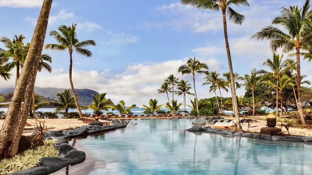 Image of a tropical resort with a large, tranquil pool surrounded by palm trees and lounge chairs. In the background, there's a sandy beach, calm ocean waters, and mountains under a partially cloudy blue sky. This idyllic setting at one of the best oceanfront hotels in Kauai exudes relaxation and natural beauty.