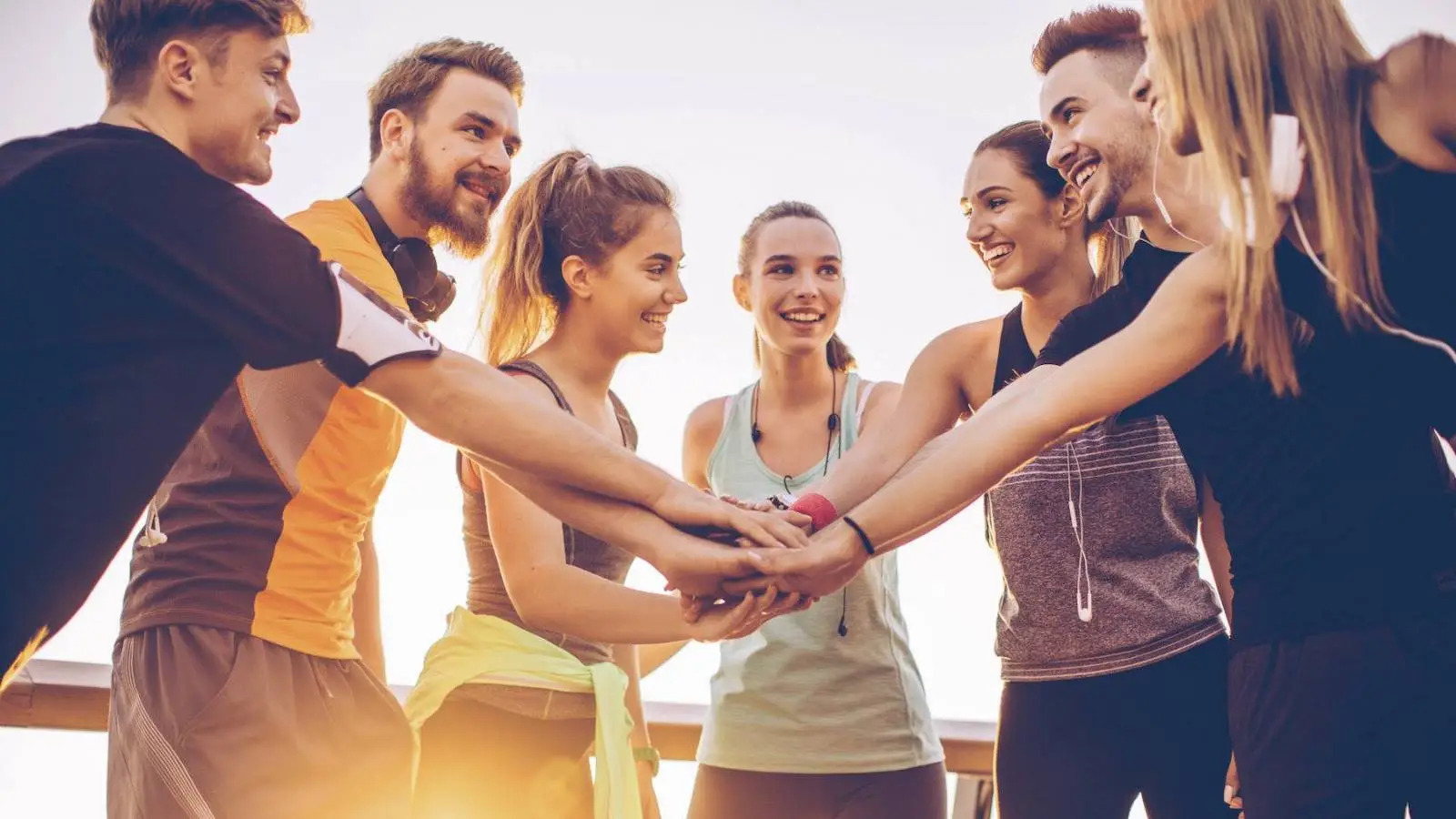 A group of eight young adults in athletic clothing stand in a circle, smiling and placing their hands together in the center. They appear to be exercising outdoors with a clear sky and sunlight in the background, enjoying one of the top family activities Sonoma County has to offer.