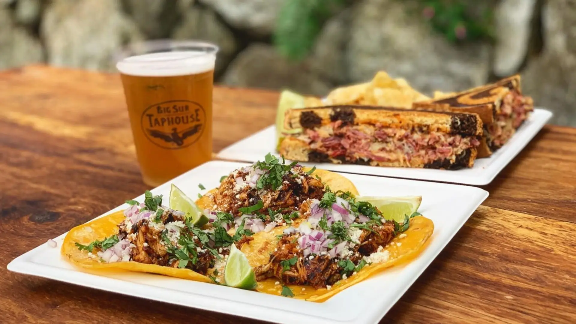 A wooden table holds a plate of three tacos filled with seasoned meat, chopped onions, cilantro, and lime wedges. Behind it, there is a pressed sandwich with a side of potato chips. A glass of beer with "Big Sur Taphouse" branding is also visible—a perfect lunch in Big Sur.