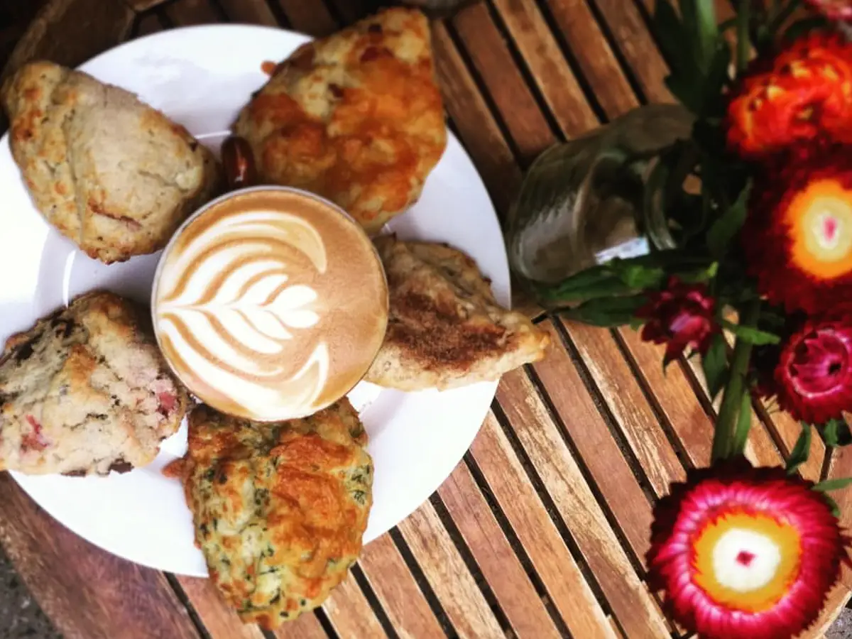 A plate of assorted pastries and a cup of coffee with latte art on a wooden table, often considered the best lunch in Santa Cruz. A vase with vibrant flowers is placed to the right of the plate. The wooden table is arranged in a striped pattern.