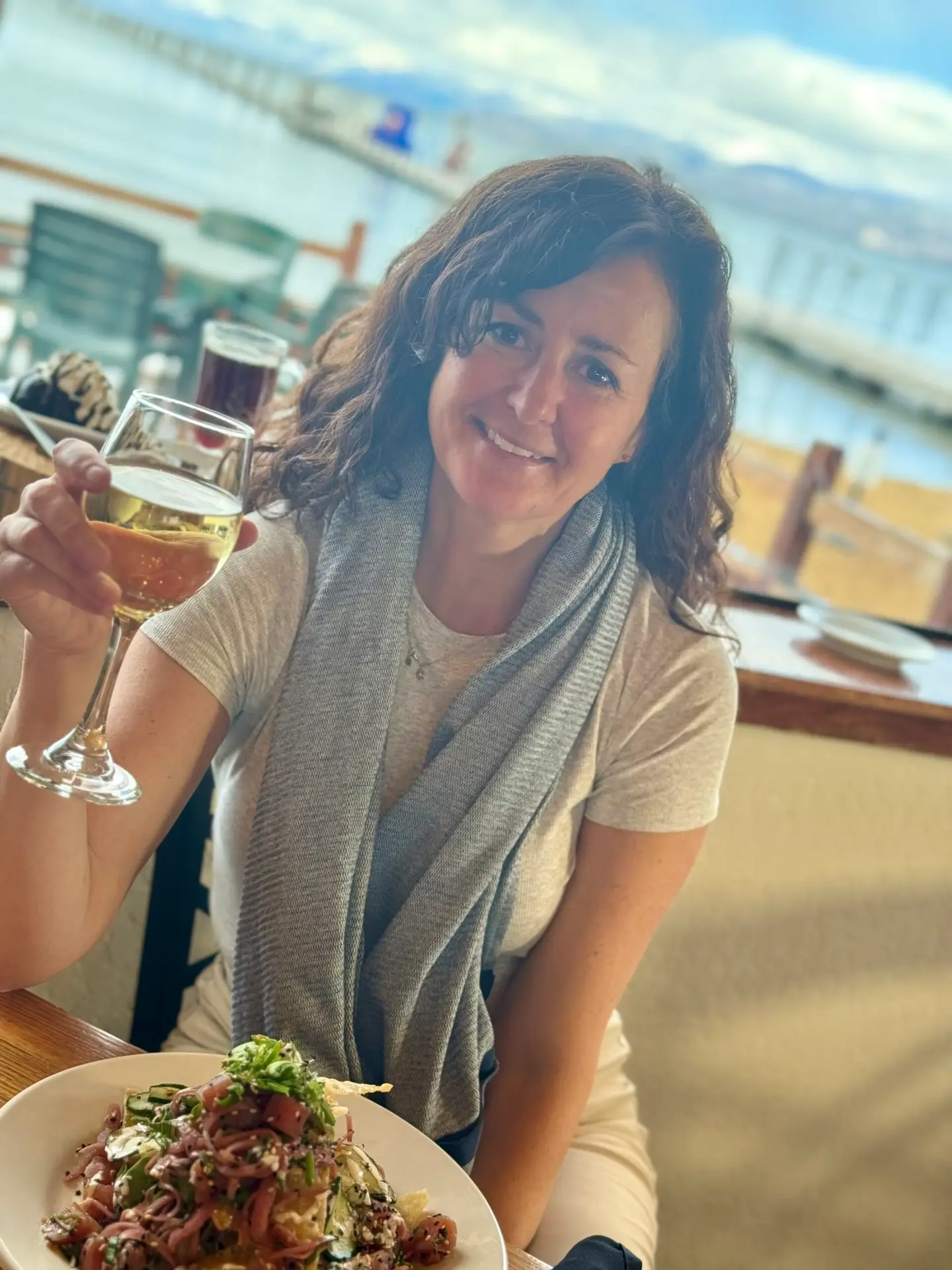 A woman with curly hair smiles while holding a glass of white wine in a restaurant. She is wearing a light-colored shirt with a grey scarf. A plate of salad is in front of her. Behind her, there is a view of a sunny beach and ocean through large windows.