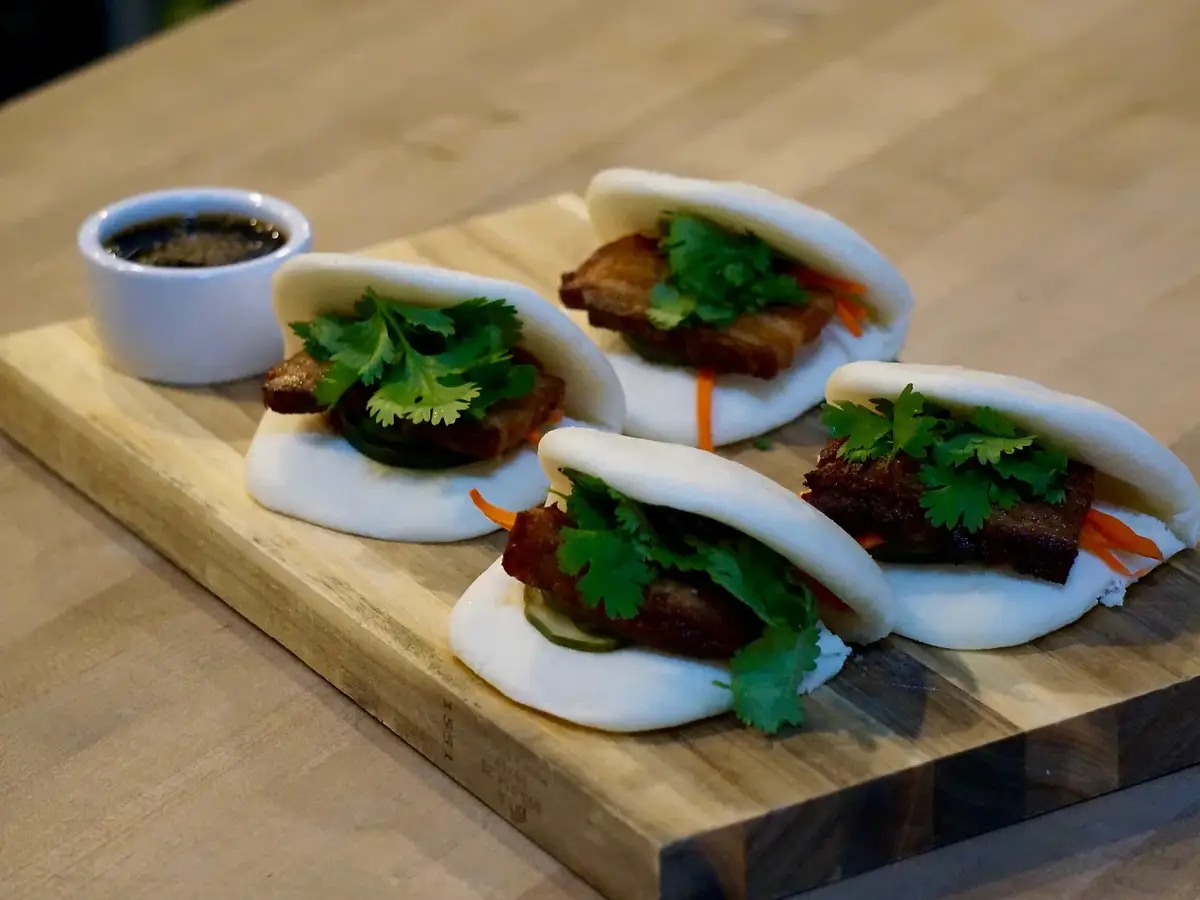 Four steamed buns filled with slices of pork belly, fresh cilantro, and pickled vegetables are arranged on a wooden board. A small white container of dipping sauce is placed nearby. Experience the best dinner in Santa Cruz with this exquisite presentation on a wooden surface.