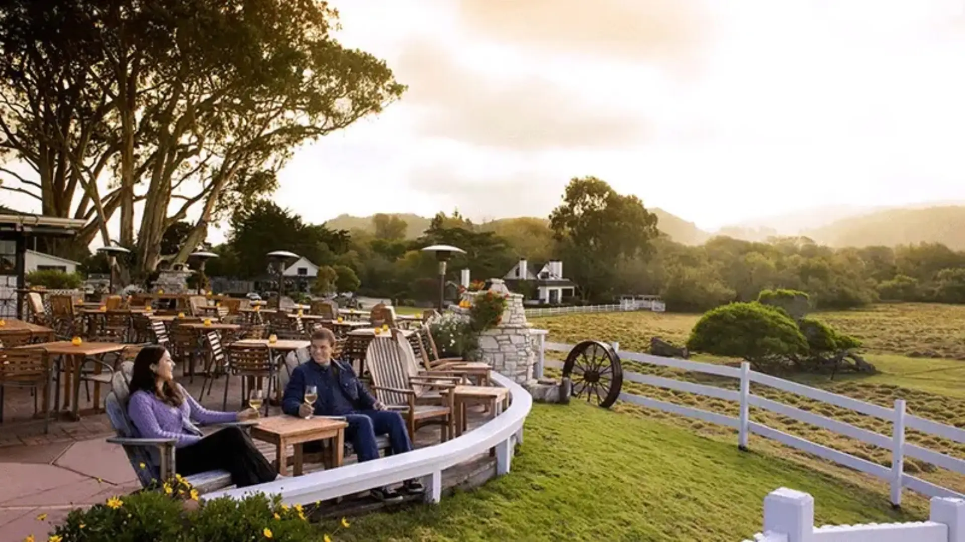 A scenic outdoor patio with wooden tables and chairs overlooks a lush green field and rolling hills, reminiscent of Ocean View Restaurants in Carmel. Two people are seated at a table, enjoying drinks. The sky is partly cloudy, and the area is surrounded by tall trees and a white fence.