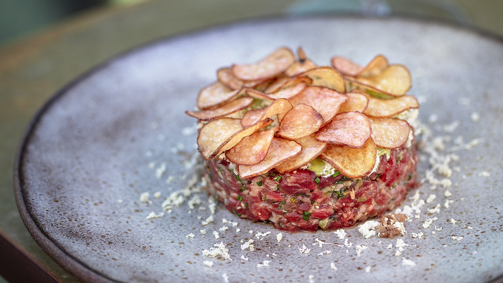 A gourmet dish of beef tartare is artfully arranged on a gray ceramic plate, topped with a generous layer of thin, crispy potato chips. Finely grated horseradish adds a hint of white color to the arrangement, reminiscent of the attention to detail found in the best seafood offerings in Big Sur.