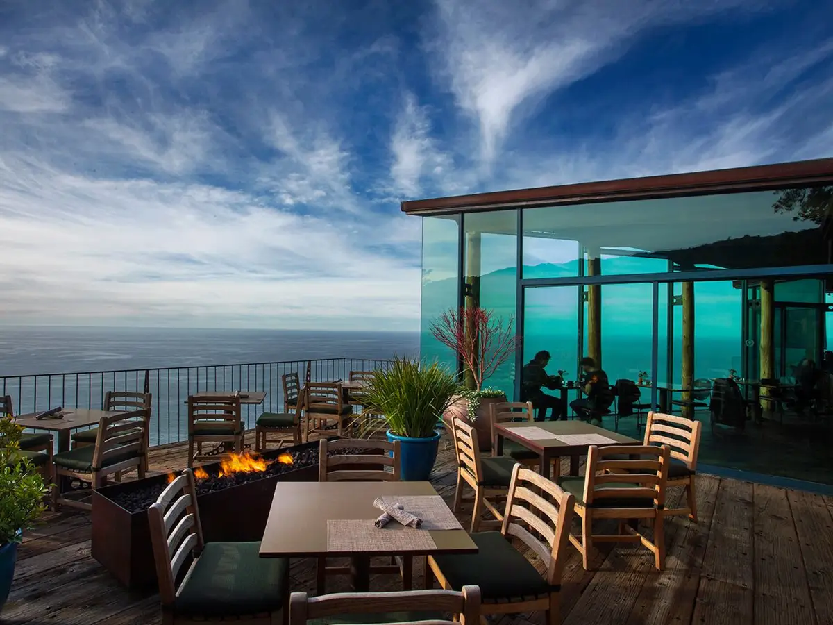 An outdoor coastal restaurant with wooden tables and chairs overlooks the ocean, offering some of the best seafood Big Sur has to offer. There's a fire pit in the center and a glass-enclosed section with more seating to the right. The sky is partly cloudy, creating a picturesque setting.