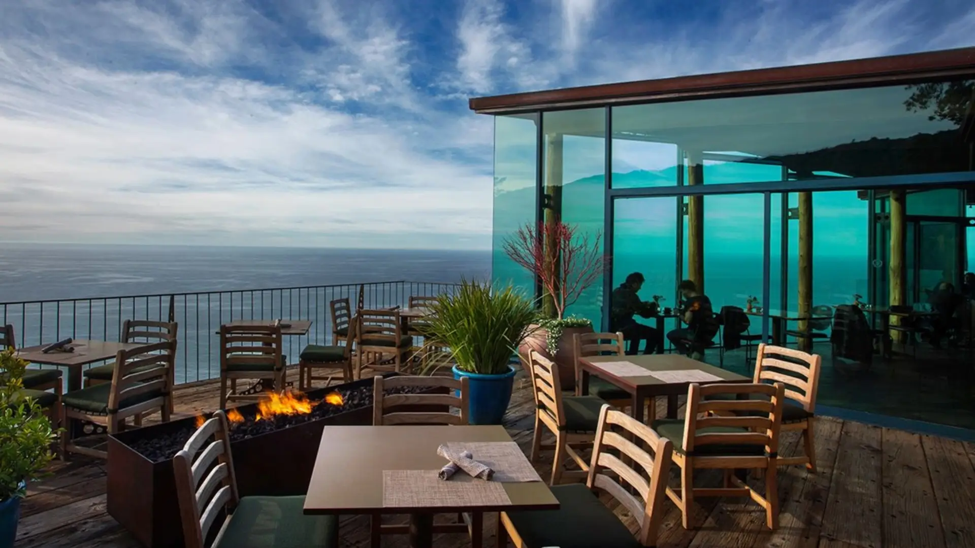 An outdoor dining area overlooks the vast ocean view of Big Sur. Wooden tables and chairs surround a fire pit on a wooden deck, with glass walls enclosing an indoor seating area. Potted plants add greenery, and the sky is partly cloudy, creating a serene atmosphere.