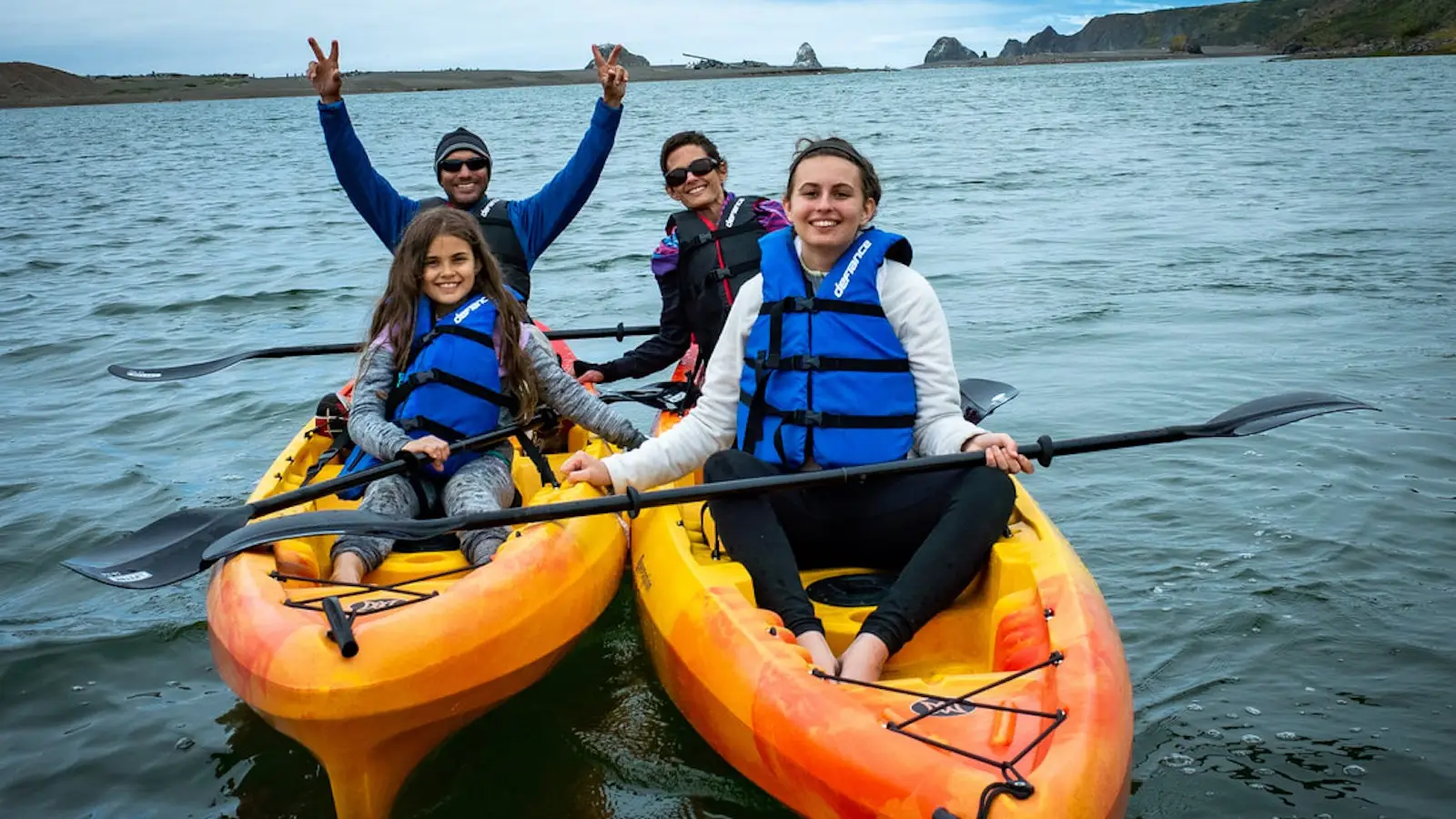 Four people enjoy a sunny day on the water while kayaking—one of the top family activities in Sonoma County. Two yellow kayaks each hold an adult and a child, all wearing blue life jackets. The adults are smiling and one is making a peace sign with both hands, while the children joyfully hold their paddles. Rocky cliffs and greenery are visible in the background.