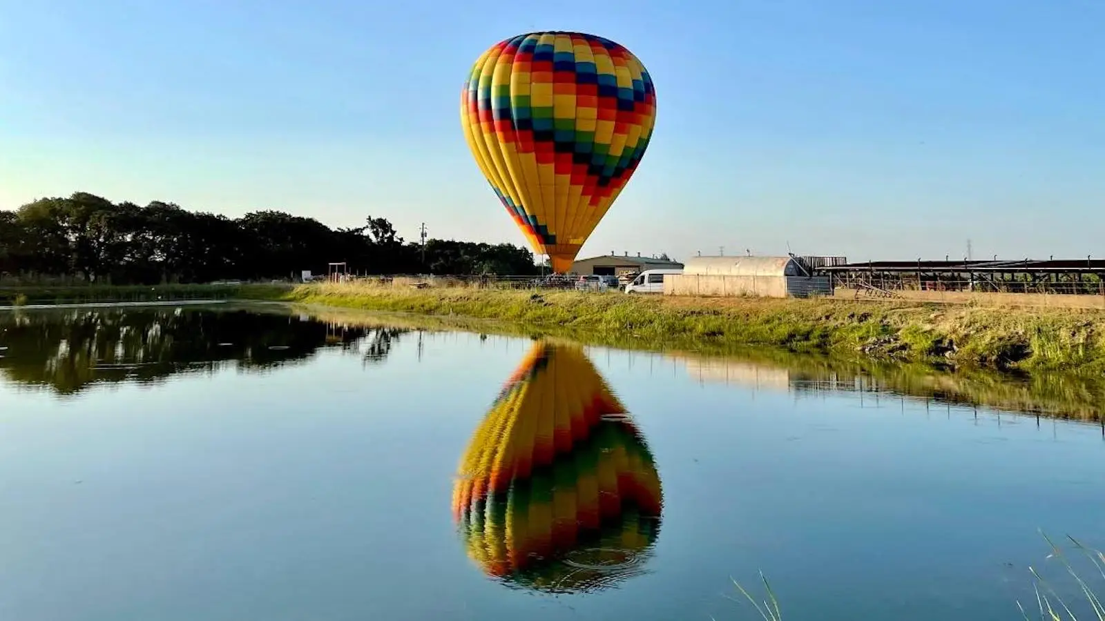 A colorful hot air balloon with a rainbow pattern floats above the ground near a body of water. The balloon's reflection is clearly visible in the calm water, and the surrounding area includes greenery and buildings under a clear blue sky—one of the top family activities Sonoma County offers.