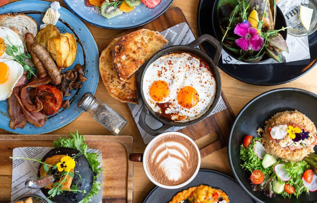 An overhead view of a table filled with various brunch dishes, including eggs and toast, a skillet with sunny-side-up eggs, a latte with latte art, a salad, and a bowl of mussels. The dishes are garnished with fresh herbs and edible flowers. Share your spread using #BrunchGiveaway for a chance to win!