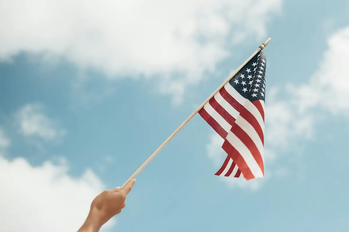 A hand holds a small American flag against a backdrop of a partly cloudy blue sky. The flag is waving slightly, with its stars and stripes clearly visible—a perfect scene to celebrate Memorial Day in the Bay Area.