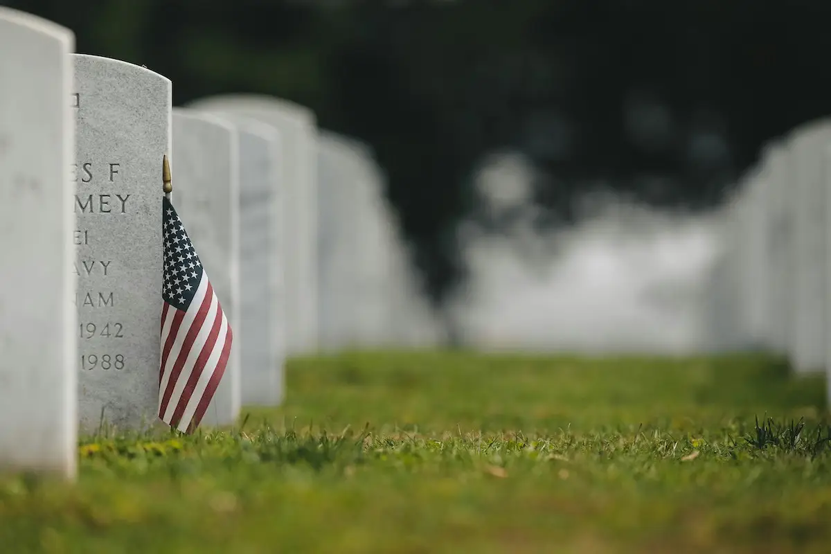 A small American flag is planted next to a white headstone in a cemetery. Other headstones can be seen in the background, out of focus. The grassy ground is well-maintained, and the overall atmosphere is solemn and respectful—a fitting tribute observed across the Bay Area on Memorial Day.