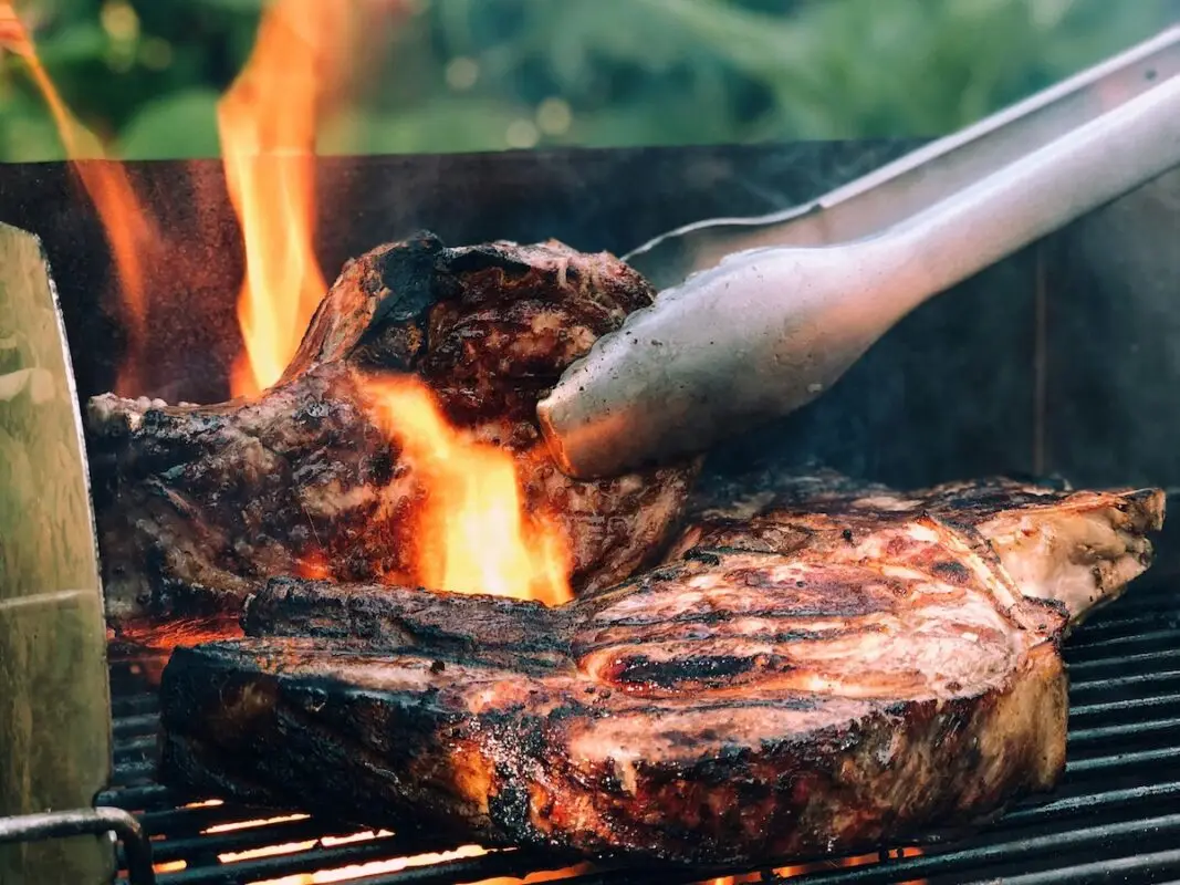 Close-up of two large, juicy steaks being grilled over an open flame during a festive Memorial Day in the Bay Area. A pair of tongs holds one of the steaks, while the fire creates a glowing orange and yellow backdrop. The steaks have noticeable grill marks and appear to be perfectly seared.