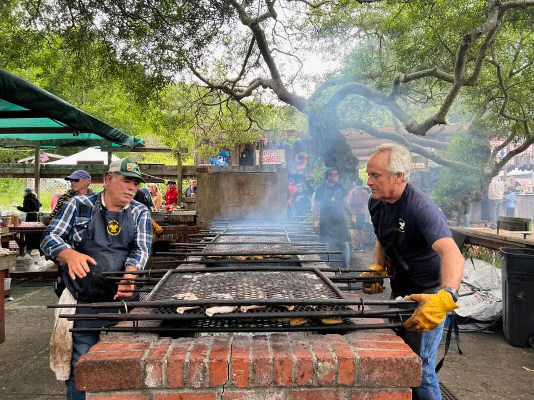 Two men cooking meat on large outdoor grills during a Memorial Day gathering in the Bay Area. One is on the left side, wearing a green cap, plaid shirt, and apron. The other, on the right, wears a dark shirt and gloves. Smoke rises from the grills, with a tree and other people in the background.