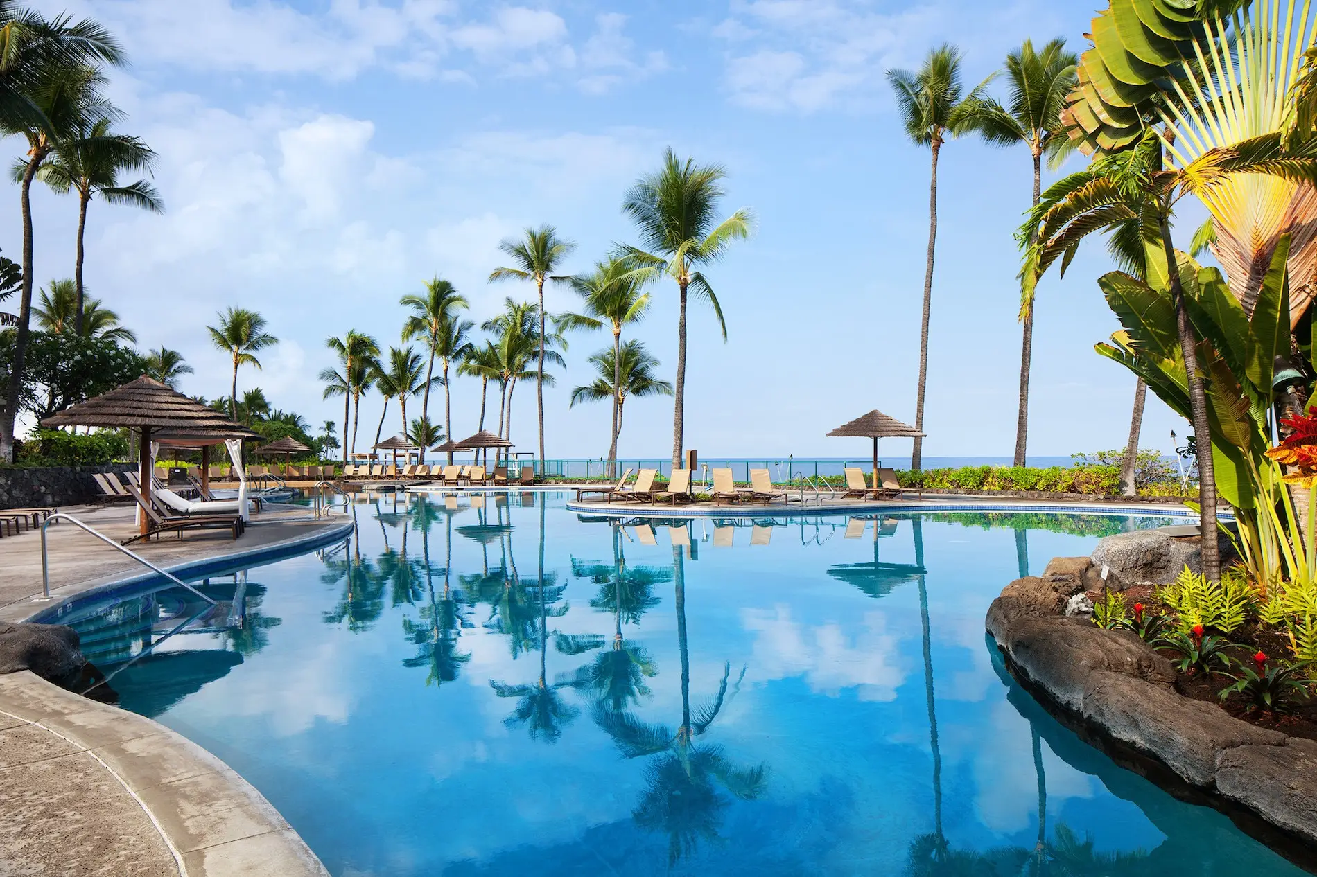 A serene tropical resort pool is surrounded by palm trees and lounge chairs. Thatched umbrellas provide shade, and the clear blue sky meets the horizon, giving a tranquil vibe. Enter our local getaways sweepstakes for a chance to experience this idyllic scenery and enhance your sense of relaxation and escape.