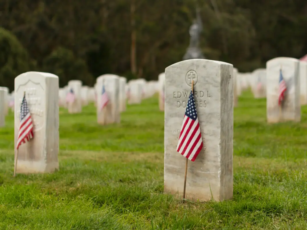 A cemetery with rows of white tombstones, each adorned with a small American flag. The grass is green and well-kept, and trees are visible in the background. The tombstones have names and dates inscribed on them, creating a poignant scene for Memorial Day in the Bay Area.