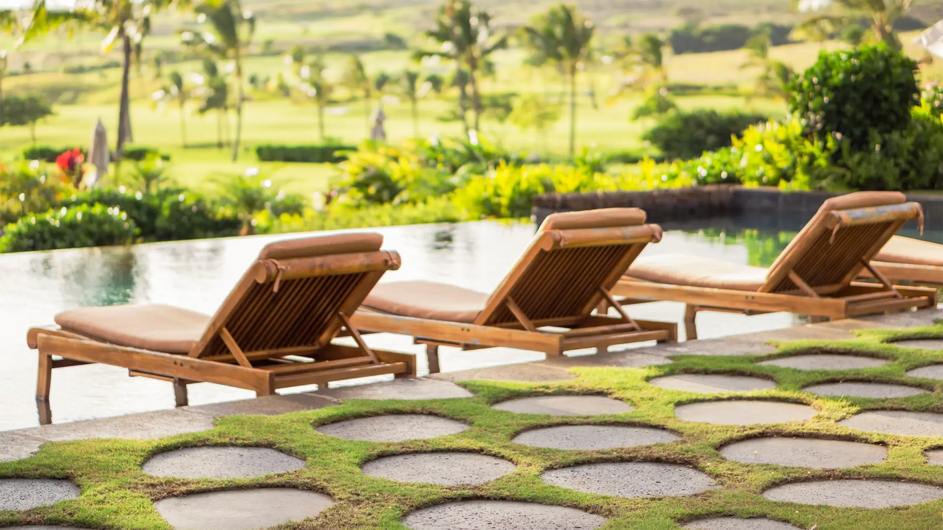 Two wooden lounge chairs with brown cushions face an infinity pool overlooking a lush, green landscape with palm trees. Circular stone pavers are set in the grass leading up to the pool area, creating a serene and relaxing outdoor environment reminiscent of the best spa hotels Kauai has to offer.