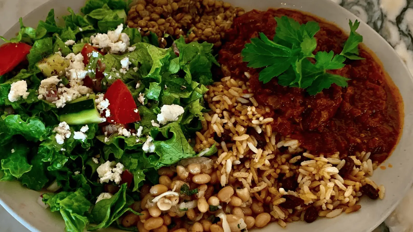 A plate of food, perfect for the best lunch on the Monterey Peninsula, featuring a mixed green salad with cherry tomatoes and crumbled cheese, a serving of rice mixed with lentils, and a portion of stewed beans garnished with fresh parsley.