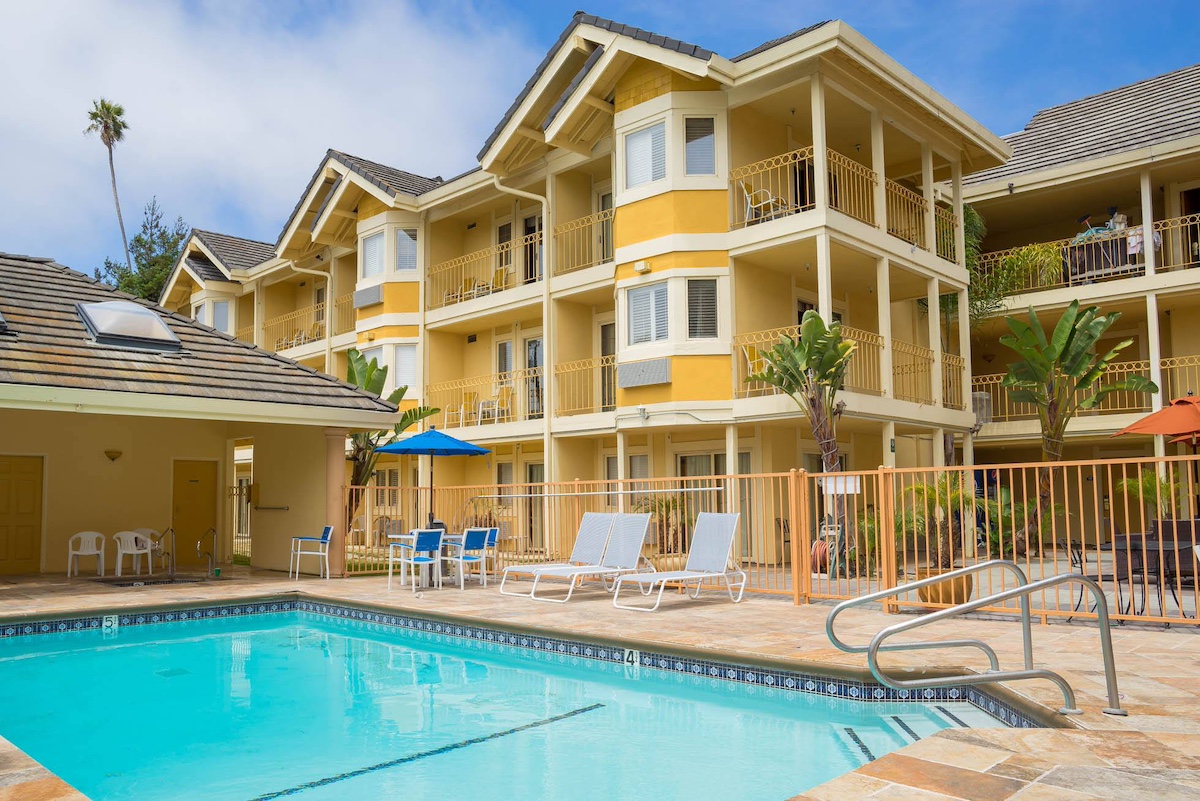 A three-story yellow apartment complex with multiple balconies is shown. In front, there is a fenced swimming pool with lounge chairs, umbrellas, and tables around it. Palm trees and a clear sky are visible in the background, making it a perfect spot for guests at Value Hotels Santa Cruz.