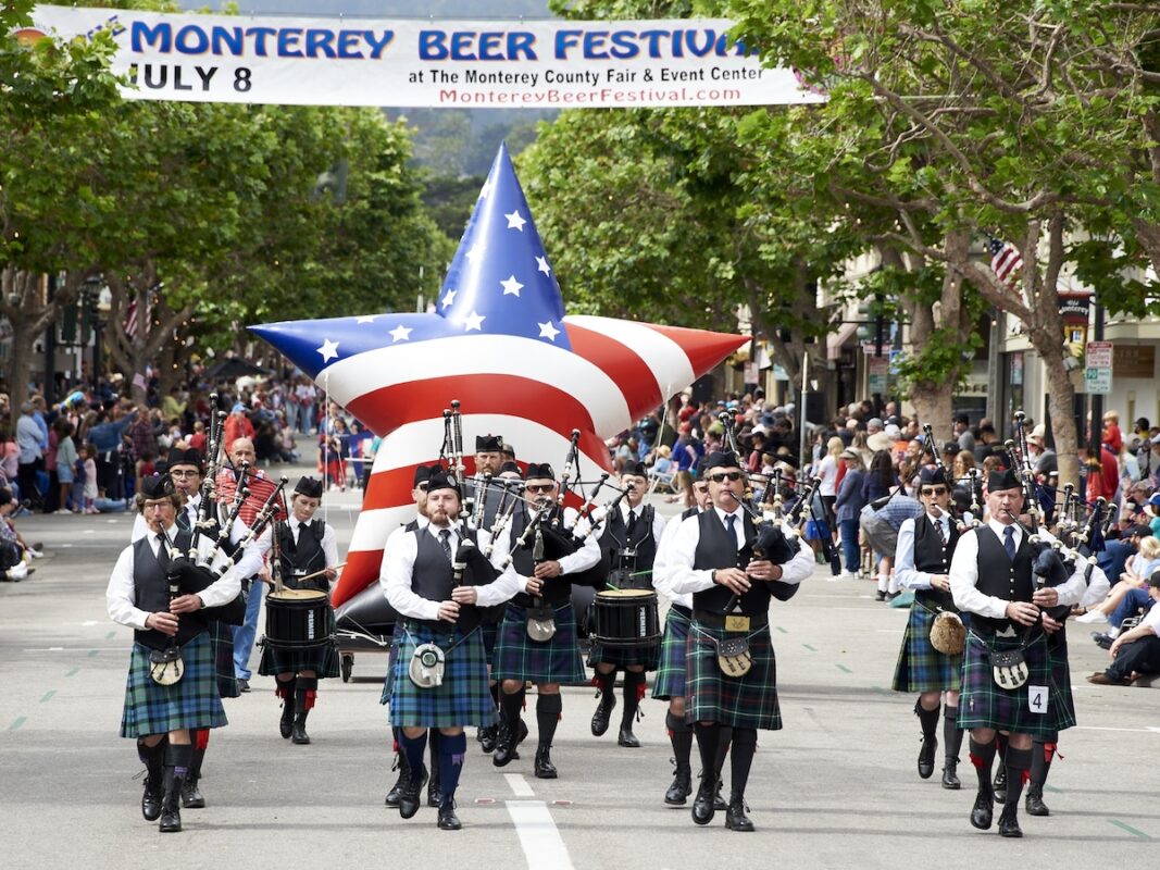 A marching band dressed in traditional Scottish attire, including kilts, parades down a street. They carry bagpipes and drums. Behind them, a large inflatable star decorated in American flag colors floats as a banner above reads "Monterey Beer Festival, July 8." One of the best things to do on the Monterey Peninsula in July.