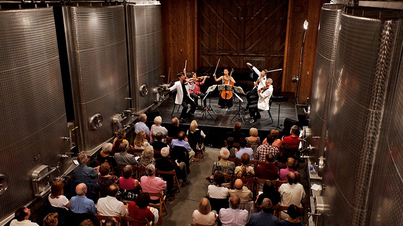 A string quartet performs on a stage in front of an attentive audience seated in rows. The setting, part of the best annual events in Napa Valley, is an industrial space with large steel tanks on either side, suggesting a winery or brewery. The musicians are fully immersed in their performance.