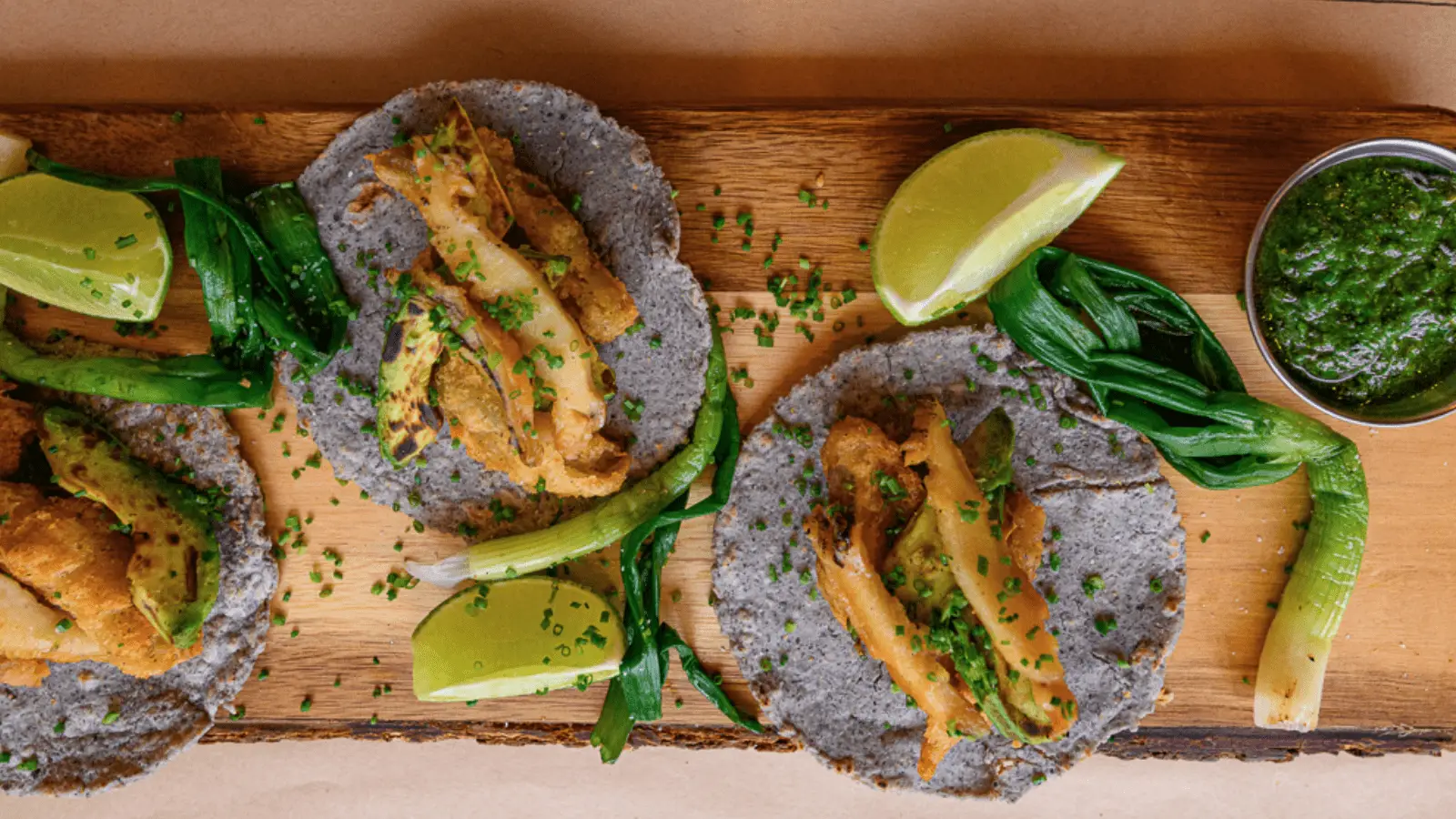 Three blue corn tacos topped with golden-brown fish, garnished with chopped chives, make for the best lunch on the Monterey Peninsula. Each taco is accompanied by lime wedges, green onions, and a small cup of green sauce, all artfully arranged on a wooden board.