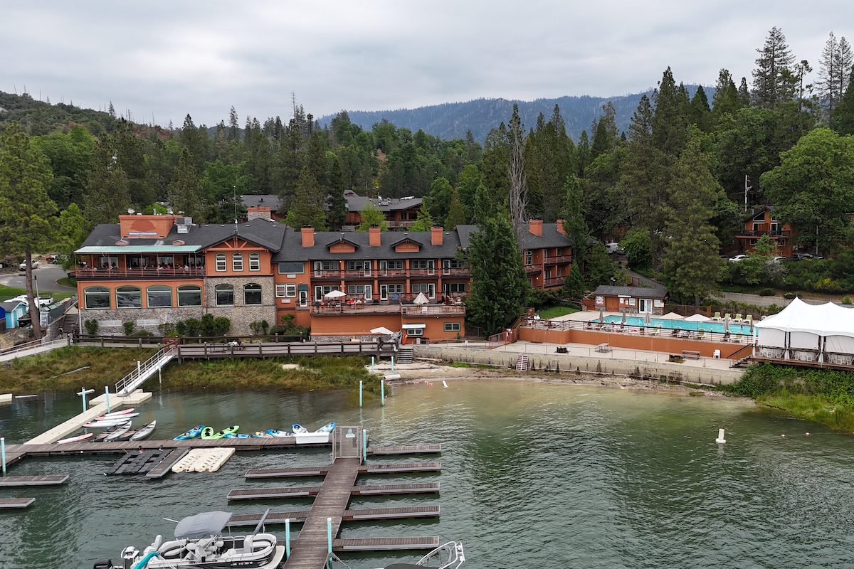 An aerial view of a lakeside resort at Bass Lake features a large building with orange accents surrounded by lush greenery and trees. It has multiple balconies and an outdoor swimming pool. The lake has several docked boats and a small beach area, perfect for a weekend getaway. Mountains are visible in the background.