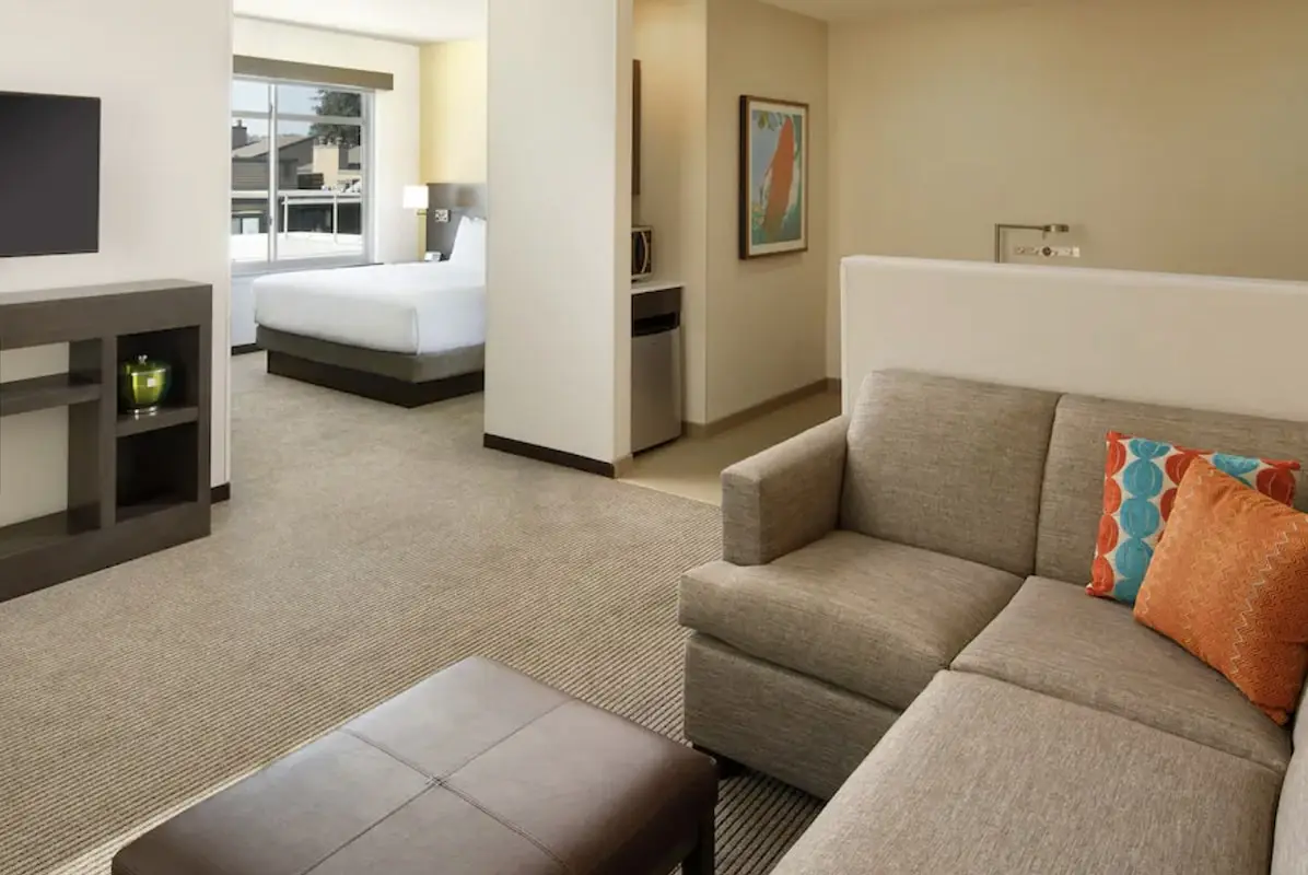 A modern hotel suite in one of the best Santa Cruz group getaway hotels features beige walls and carpet. There is a gray sofa with bright orange and blue decorative pillows in the living area, a dark brown ottoman, and a wall-mounted TV. In the background is a sleeping area with a bed and large window.