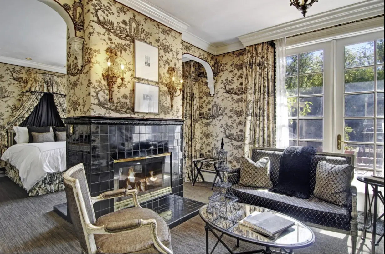 Image of a luxurious living room and bedroom combination with elegant black and white decor, reminiscent of Santa Cruz romantic hotels. The space features a tiled fireplace, ornate wall patterns, stylish furniture including a sofa and chairs, and large windows letting in natural light.