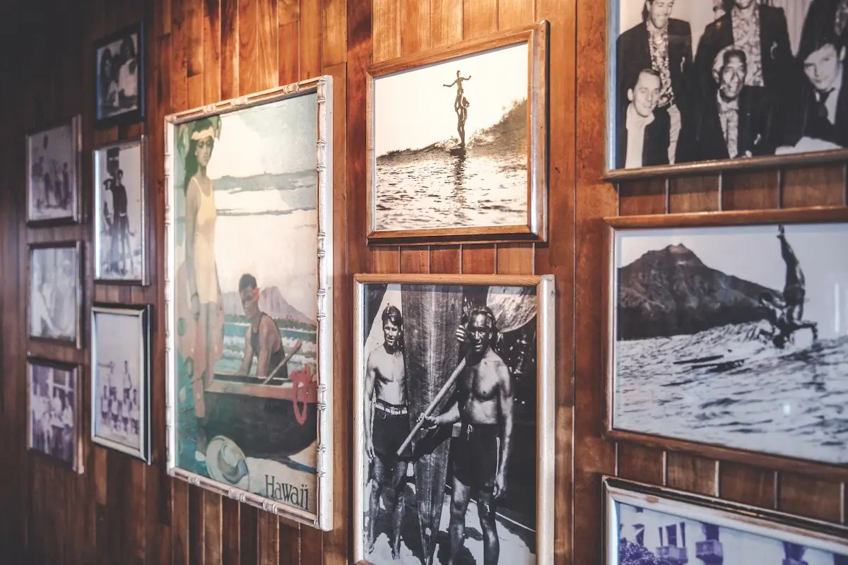 A wooden-paneled wall at Duke's Restaurant adorned with a variety of framed vintage photographs. The images depict people surfing, beach scenes, and moments from mid-20th century Hawaii. Some photos are black and white, while others are in faded color.