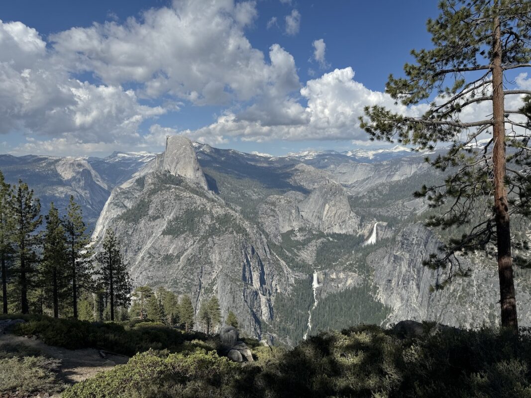 A scenic view of Yosemite National Park with granite peaks, including the prominent Half Dome in the center. Waterfalls cascade down cliffs in the distance. Pine trees frame the foreground, and the sky is partly cloudy—a perfect setting for a Bass Lake weekend escape.