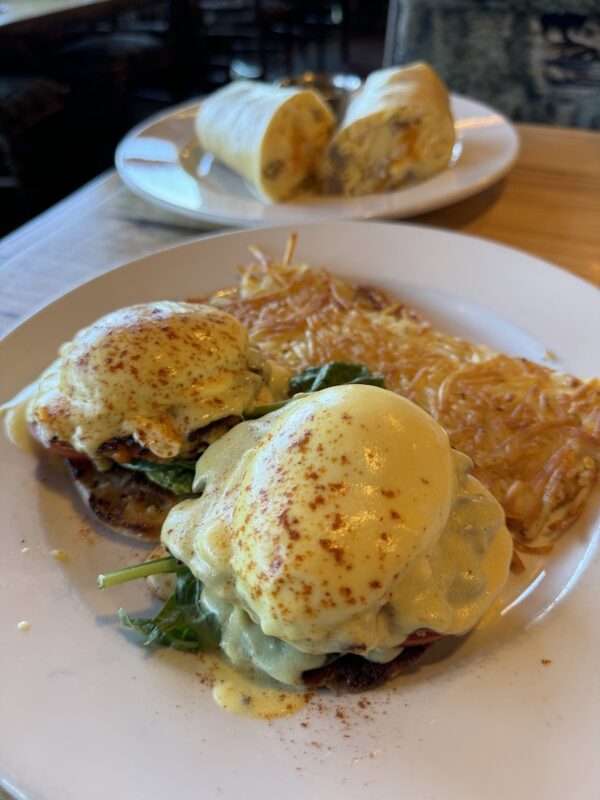 A plate of eggs Benedict, topped with hollandaise sauce and sprinkled with paprika, accompanied by a side of hash browns. In the background, there is a burrito cut in half on a separate plate. Both dishes are set on a wooden table, capturing the essence of a Bass Lake weekend breakfast.