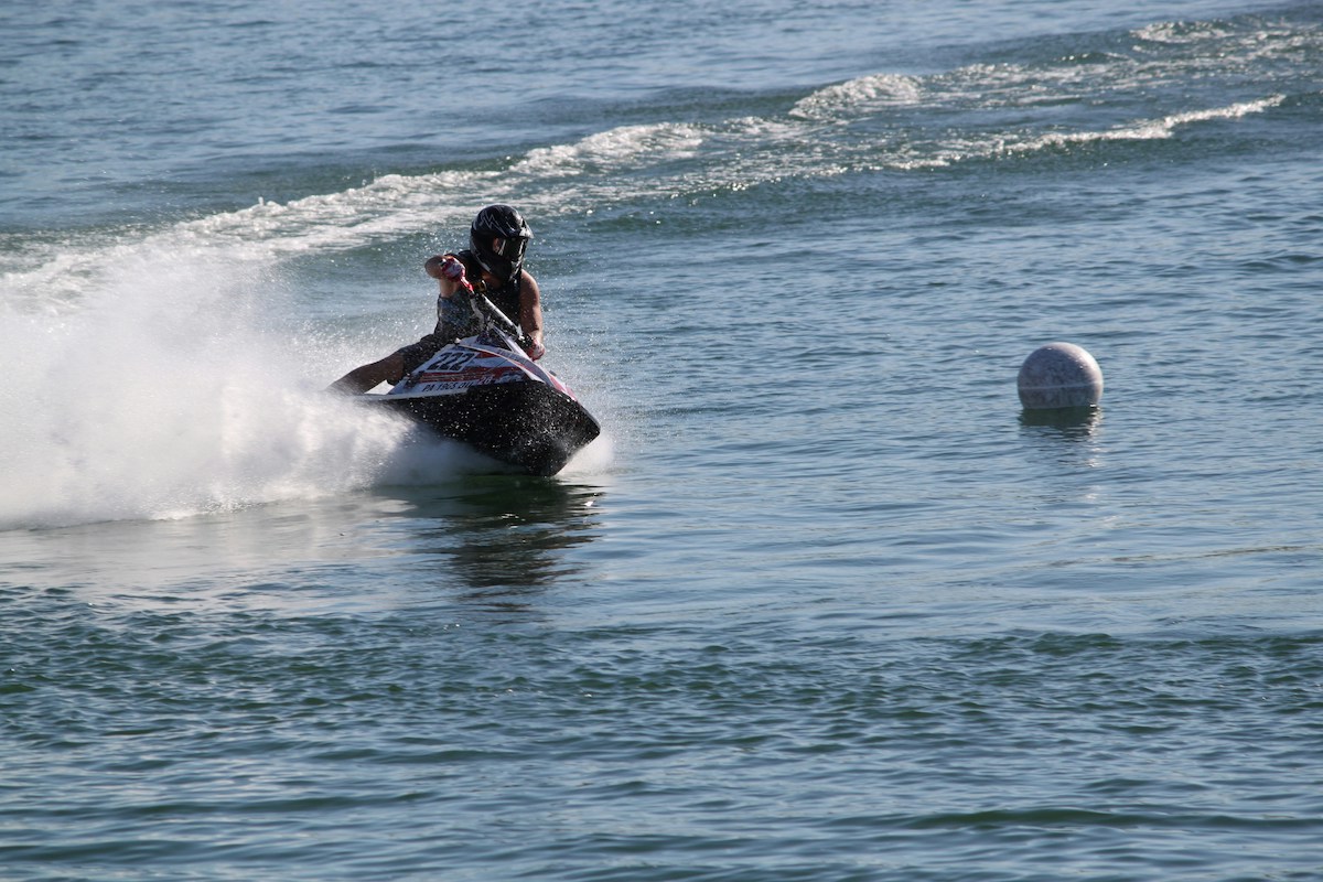 A person wearing a helmet rides a jet ski at high speed on Bass Lake, creating a spray of water behind. The water surface shows ripples, and there is a buoy floating nearby. The background features a wide expanse of water under a clear sky, perfect for an adrenaline-filled weekend.